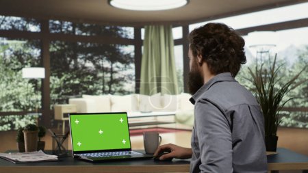 Foto de Rico inversor se relaja en su elegante chalet de montaña mientras mira la pantalla verde en su portátil. Supervisor extranjero trabaja con panel de maqueta aislado en el ordenador portátil en la vida próspera. - Imagen libre de derechos