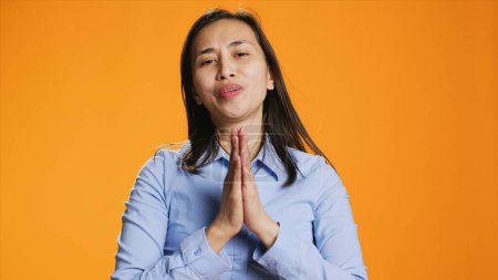 Photo for Filipino model praying for forgiveness and showing gratitude on camera, hopeful woman with belief in spirituality. Asian person with prayer hands having faith in God, orange background. - Royalty Free Image