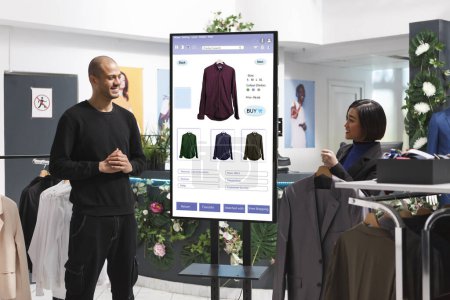 Photo for Male customer is shown clothing on an interactive kiosk board by female assistant in mall clothing store. Asian retail employee assisting a client who is self-ordering modern fashion items. - Royalty Free Image