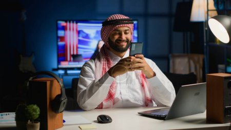 Photo for Arabic man at home in living room typing messages on smartphone. Muslim person holding mobile phone, enjoying relaxing leisure time talking with friends over online messaging app - Royalty Free Image