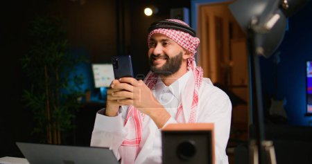 Photo for Portrait of happy arabic teleworker sending text messages to friends while working from home in stylish apartment. Cheerful Middle Eastern man typing on mobile phone with opened tv as background noise - Royalty Free Image