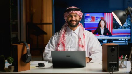 Photo for Portrait of smiling Arab businessman working at modern desk, typing on his laptop, solving tasks. Muslim guy browsing on digital device, doing email communication in professional office setting - Royalty Free Image