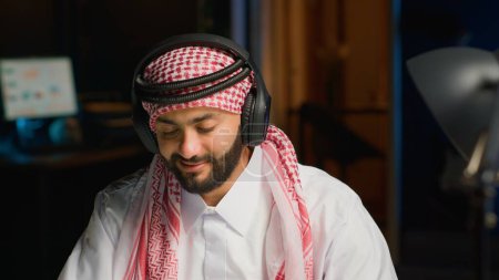 Téléchargez les photos : Employé arabe avec casque sans fil enthousiaste à l'idée de travailler depuis le bureau de l'appartement. Entrepreneur musulman faisant des recherches en ligne tout en écoutant de la musique pour faire passer le temps plus rapidement, gros plan - en image libre de droit