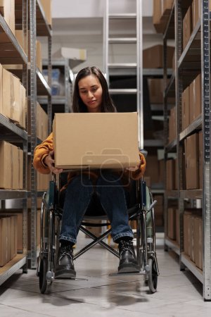 Photo for Warehouse asian worker in wheelchair holding packed parcel ready to distribution. Storehouse asian woman employee with disability working in storage room aisle with shelves full of cardboard boxes - Royalty Free Image