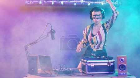 Photo for Cool DJ artist using vinyl records to mix music at turntables, doing remix mash of techno tunes using electronics. Performer mixing with stereo mixer, doing performance at party. Tripod shot. - Royalty Free Image