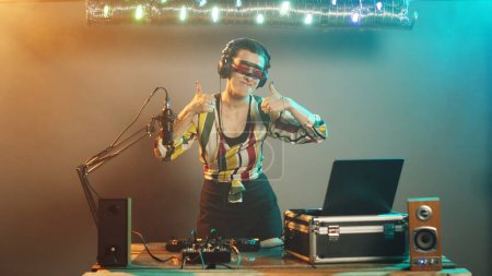 Photo for Disc jockey gives thumbs up while mixing music at techno party, creating funky atmosphere on stage at club. Woman DJ using bass control buttons and stereo equipment for remix. Tripod shot. - Royalty Free Image