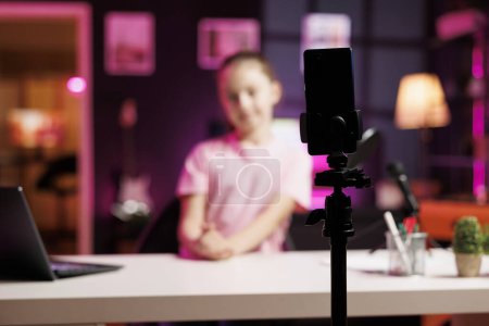 Photo for Young child in blurry background doing livestream using smartphone on professional recording tripod. Cute kid does live broadcast using cellphone camera to talk with online audience, focus on phone - Royalty Free Image
