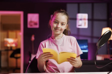 Photo for Kid influencer in studio reading newly released hardcover book from publisher. Cute girl filming literature video essay, presenting new novel from author to readers in pink neon lit studio - Royalty Free Image