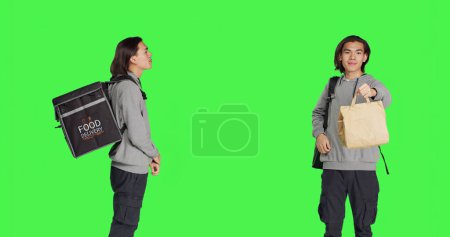 Photo for Standing over studio greenscreen backdrop, asian man operates on food delivery with heated bag. Young adult holding backpack for takeout, prepared to give ordered meals to clients. - Royalty Free Image