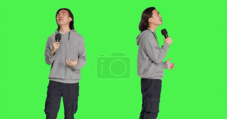 Photo for Trendy guy performs karaoke on camera, using recording device to sing popular melodies and lyrical content. Young man having an excellent time over greenscreen backdrop, creating sounds and songs. - Royalty Free Image