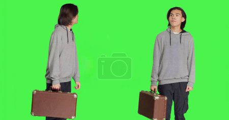 Photo for Young person carries vintage briefcase over greenscreen template in studio, waiting impatiently for something. Asian guy holds big suitcase while he checks time on wrist watch. - Royalty Free Image