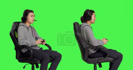 Photo for Cheerful person enjoys online gameplay, sitting on gaming chair over greenscreen isolated background in studio. Young asian guy having fun with competition, man playing video games. - Royalty Free Image