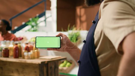 Photo for Local vendor holds greenscreen phone with isolated display, presenting copyspace technology on mobile phone. Person looking at blank chromakey screen at grocery store register. - Royalty Free Image