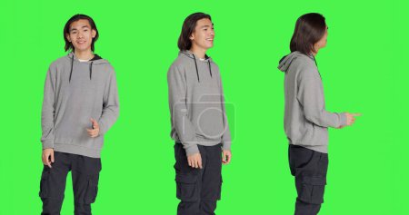 Photo for Asian person shows approval sign over greenscreen backdrop, doing yes gesture and smiling. Young man with optimistic facial expressions feeling pleased and expressing agreement. - Royalty Free Image
