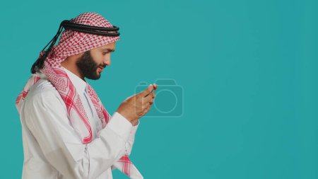 Photo for Joyful muslim man having fun with online game on his smartphone, wearing traditional islamic costume. In the studio, arab adult enjoying e sport challenge with mobile phone device. - Royalty Free Image