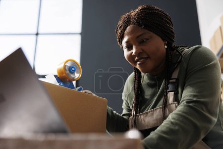 Photo for African american worker checking clients orders on laptop computer, preparing packages in warehouse. Stockroom supervisor using cardboard boxes for products shipping in storage room - Royalty Free Image
