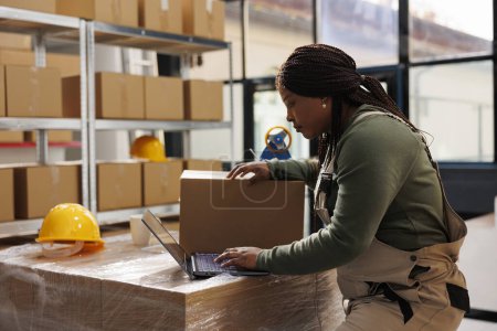Photo for African american worker checking customers orders on laptop, preparing packages using cardboard boxes in storage room. Warehouse manager wearing industrial overall during inventory - Royalty Free Image