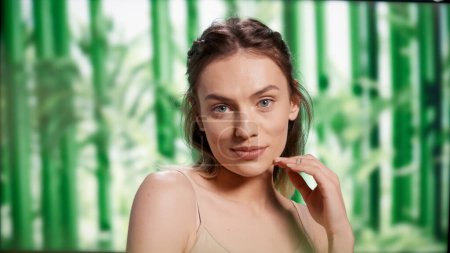 Téléchargez les photos : Smiling flawless woman with bare luminous skin posing on camera, standing over background with bamboo trees. Happy beautiful girl promoting cosmetics and skincare routine in studio. - en image libre de droit