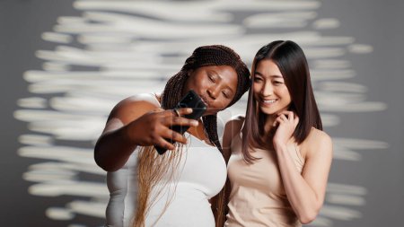 Téléchargez les photos : Two skincare models taking photos with smartphone, diverse young women advertising different skintones and body shapes. Beautiful luminous girls having fun with pictures for beauty ad. - en image libre de droit