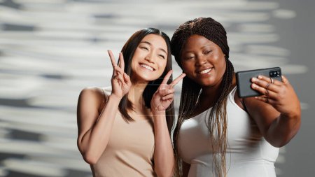 Foto de Interracial confident girls taking pictures on smartphone, having fun with photos on mobile phone in studio. Happy funny models being positive and posing for body confidence campaign. - Imagen libre de derechos