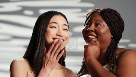 Téléchargez les photos : African american woman sharing secret with asian girl, creating skincare and self love campaign. Advertising body positivity and self acceptance, interracial friends laughing. Beautiful girls. - en image libre de droit