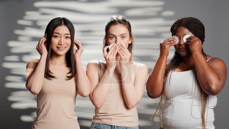 Foto de Young models doing three wise monkeys sign with cotton pads, creating beauty skincare campaign in studio. Diverse women having fun playing around with cosmetics, cleansing skin. - Imagen libre de derechos