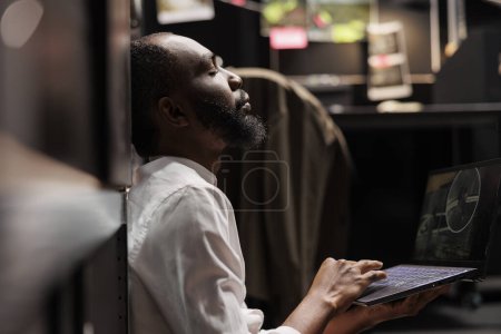 Photo for Tired police agent sitting with closed eyes, solving complex crime case in dark office. Exhausted private detective spying on suspect, watching cctv camera records on laptop - Royalty Free Image