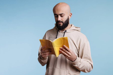 Photo for Young thoughtful focused arab man reading novel from book with yellow cover. Concentrated bald bearded person holding open softcover textbook and studying with pensive expression - Royalty Free Image