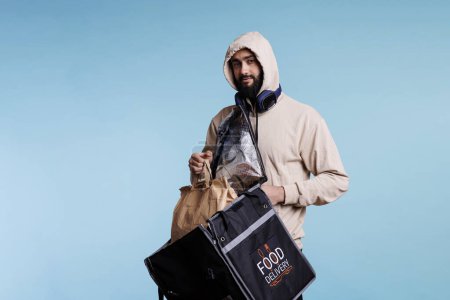 Téléchargez les photos : Jeune homme arabe prenant sac en papier avec de la nourriture du sac à dos et regardant la caméra. Livreur dans le capot tenant sac thermique et paquet avec déjeuner à emporter portrait studio sur fond bleu - en image libre de droit