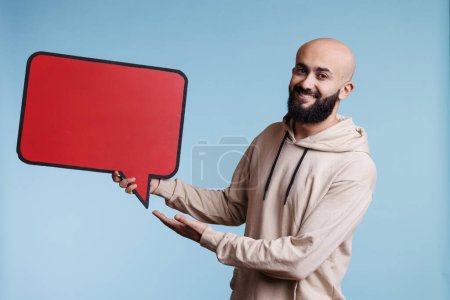Photo for Cheerful arab man promoting new product with comic balloon red banner portrait. Smiling person holding empty dialog bubble and looking at camera with happy facial expression - Royalty Free Image