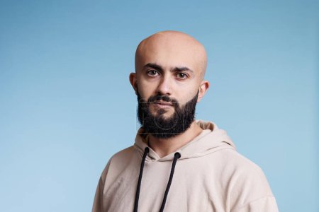 Photo for Young bald arab man with relaxed facial expression studio portrait. Bearded arabian person with neutral emotions wearing casual beige hoodie and looking at camera on blue background - Royalty Free Image