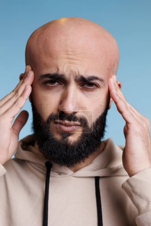 Foto de Hombre árabe sintiendo dolor en la cabeza mientras frota las sienes y mira a la cámara. Joven guapo que sufre de dolor de cabeza con exhausto estudio de expresión facial retrato de cerca - Imagen libre de derechos