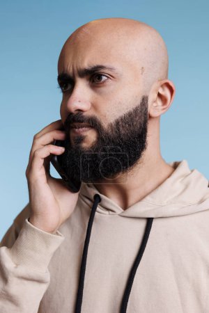 Photo for Serious arab man talking on smartphone and looking away. Young bald bearded arabian person answering mobile phone call and listening to interlocutor with tensed facial expression - Royalty Free Image