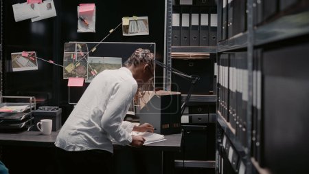 Foto de Woman detective uncovering clues on evidence board, working on crime solving in archive room. Female investigator examining surveillance footage, searching for insight and details. - Imagen libre de derechos