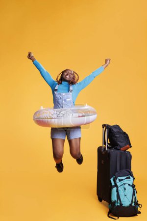 Foto de Mujer alegre viajando al extranjero con bolsas, sintiéndose emocionada por las vacaciones internacionales en el estudio. Viajero femenino saltando alrededor con inflable, siendo tonto que sale en viaje de vacaciones. - Imagen libre de derechos