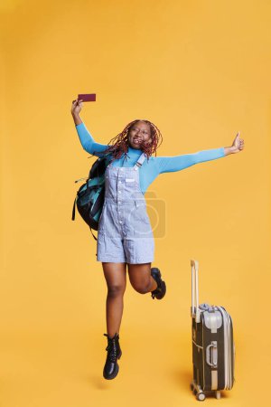 Foto de Chica sonriente saltando en cámara, sosteniendo el pase de identificación y llevando bolsas de equipaje. Joven turista emocionada con destino internacional, vacaciones con pasaporte. - Imagen libre de derechos