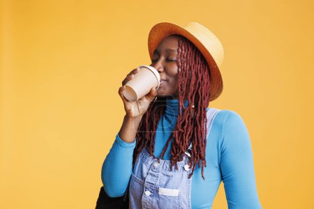 Téléchargez les photos : Femme afro-américaine dégustant une tasse de café en studio, se sentant joyeuse avant une escapade internationale. Jeune voyageuse buvant de la caféine et s'amusant, étant heureuse à la caméra. - en image libre de droit