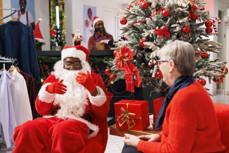 Téléchargez les photos : Employé du Père Noël donne des cadeaux à la femme âgée en magasin, offrant boîte festive à côté de l'arbre de Noël au centre commercial. Personne âgée se sentant gaie après avoir reçu des marchandises gratuitement, ventes saisonnières. - en image libre de droit