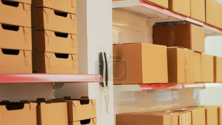 Photo for Empty warehouse with landline phone on wall, shelves and racks filled with merchandise in cardboard boxes. Multiple products prepared for shipment and transportation, stock goods. - Royalty Free Image