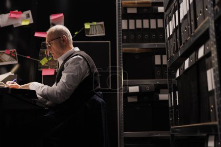 Photo for Elderly investigator working overtime at crime scene report, analyzing evidence files in arhive room. Overworked private detective checking criminology documents, planning strategy to catch suspect - Royalty Free Image