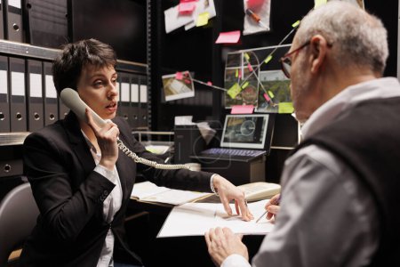 Photo for Private detective talking with remote victim at landline phone, discussing mysterious suspect profile. Criminology team working late ar night at criminal case, analyzing crime scene evidence - Royalty Free Image