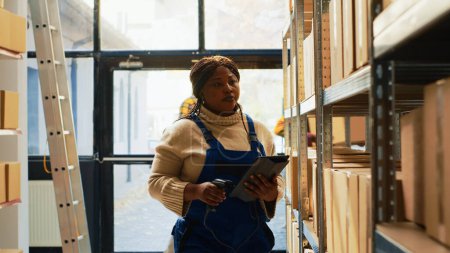 Foto de Mujer afroamericana escaneando códigos de barras en cajas, trabajando con escáner y tableta para revisar mercancía o estantes y estantes. Empleada mirando productos al por menor para inventario de depósito. - Imagen libre de derechos
