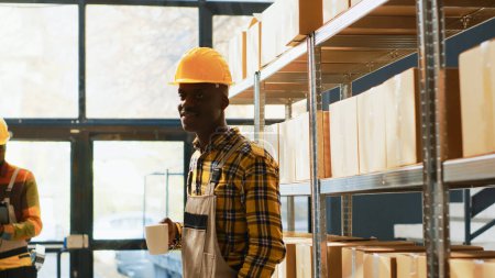 Foto de African american warehouse worker posing near shelves filled with products in boxes. Young man with overalls and helmet working in storage room depot with retail store merchandise. - Imagen libre de derechos
