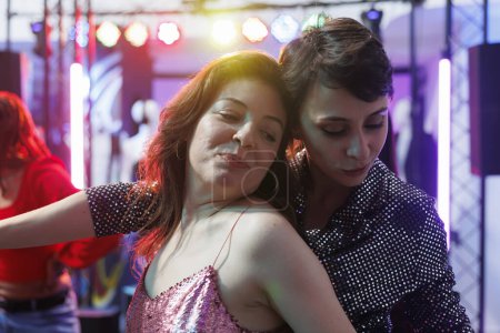 Photo for Smiling women hugging while dancing together at nightclub party. Young romantic girlfriends couple relaxing and moving on crowded dancefloor while clubbing at discotheque - Royalty Free Image