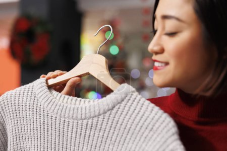 Photo for Buyer analyzing clothes on hangers before buying gifts for christmas eve celebration, woman looking at sweater fabric. Asian client checking new sales merchandise in store. Close up. - Royalty Free Image