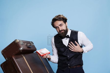 Photo for Hotel concierge holds cardiology drugs for heart problems, wearing formal attire in front of camera. Doorman with heartache recommending box of pills or supplements as medication. - Royalty Free Image