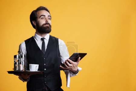 Photo for Luxury butler checking list of reservations on tablet, carrying tray with food and drinks. Elegant restaurant waiter preparing to give bill or tab to customers, using modern device. - Royalty Free Image