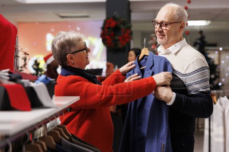 Photo for Senior couple examining blazers on racks in mall clothing store, looking for formal attire to wear on christmas dinner festivity. Elderly customers searching for elegant clothes in festive shop. - Royalty Free Image