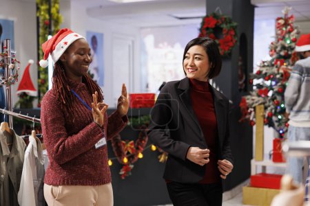 Photo for Asian woman trying on formal clothes before buying on sale, assistant helping customer to choose items for festive christmas event with family. African american employee showing jacket to person. - Royalty Free Image