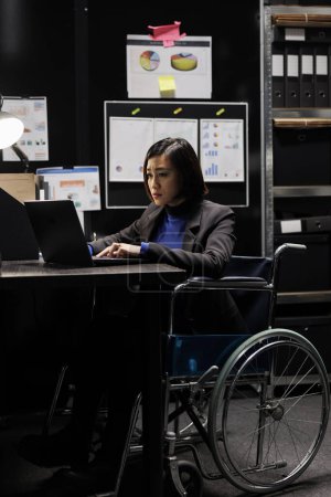 Photo for Paralyzed businesswoman accountancy executive in wheelchair adding budget plan data on laptop. Asian woman with paraplegia in storage office filled with paperwork folders on shelves and flowcharts - Royalty Free Image